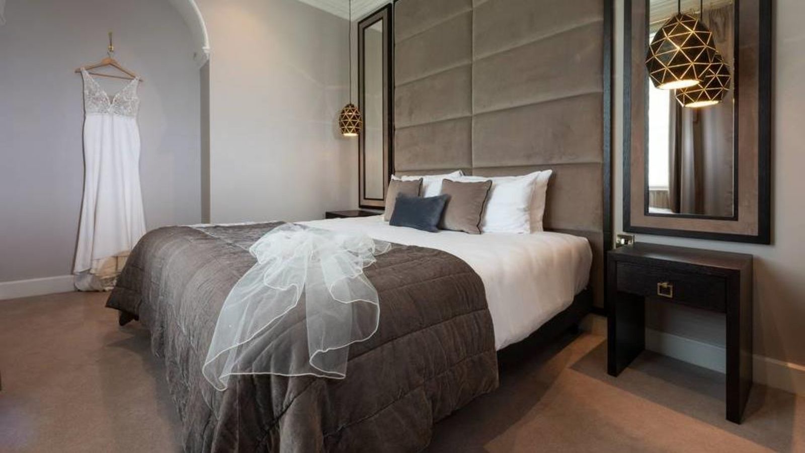 A suite with a grey upholstered headboard and a wedding dress hanging by the window