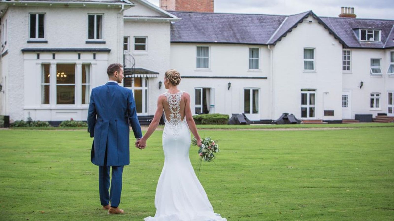 A bride and groom walking hand in hand across the lawn towards the hotel